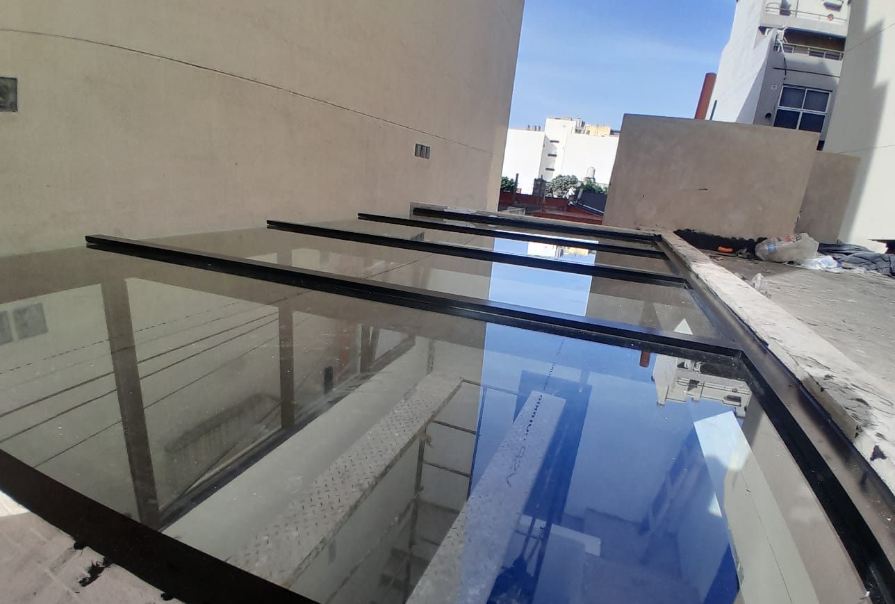 Techo de vidrio con perfiles de aluminio negros que refleja el cielo entre edificios
